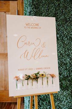 a welcome sign with flowers on it in front of a green wall and wooden easel