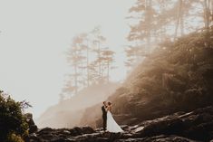 a bride and groom standing on rocks in the woods at their wedding day with sun shining through the trees