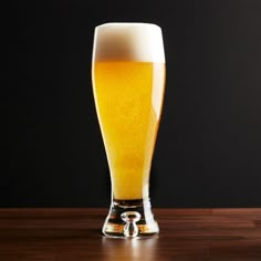 a tall glass filled with beer sitting on top of a wooden table next to a black wall