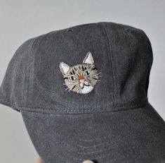 a gray hat with a cat embroidered on it's front and the face of a cat