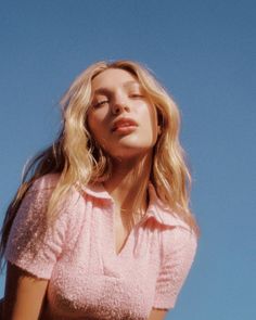 a woman with her eyes closed standing in front of a blue sky wearing a pink top