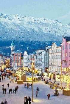 people are walking around in the town square with christmas lights on trees and snow covered mountains behind them