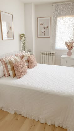 a white bed with pink and tan pillows in a bedroom next to a window on a hard wood floor