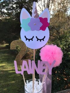 a cupcake topper with a unicorn face and pink pom - poms