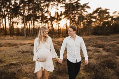 a pregnant couple holding hands and walking through the grass in front of some tall trees