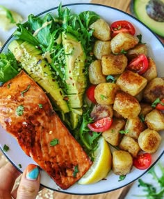 a person holding a plate with salmon, potatoes and avocado on the side