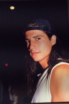 a man with long hair wearing a baseball cap and white tank top looking at the camera