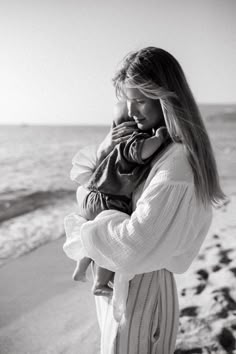 a woman standing on the beach holding a baby in her arms and talking on a cell phone