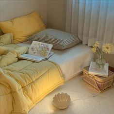 a bed sitting next to a window covered in yellow sheets and pillows with a book on top of it