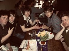 four men sitting at a table with food and drinks in front of them, posing for the camera