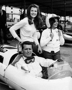 two women and a man sitting in a race car with the driver standing behind them