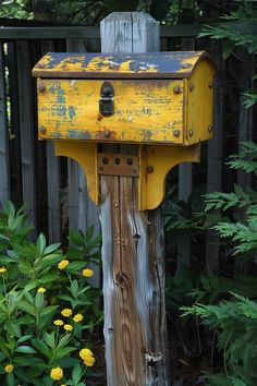 an old yellow mailbox sitting on top of a wooden post next to some flowers