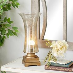 a table with a mirror, vase and flowers on it next to a lamp that is turned on