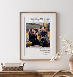 two women sitting next to each other in front of a white wall with a framed photo