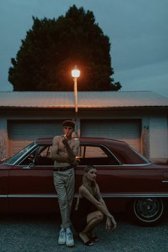 a man and woman standing next to a red car at night with a street light in the background