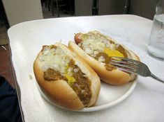 two hotdogs with chili and cheese on a white plate next to a fork