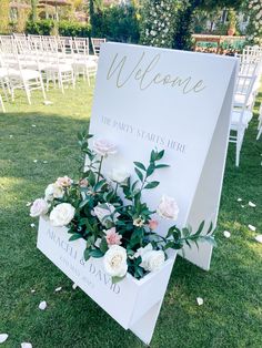 a welcome sign with flowers and greenery on the grass in front of an outdoor ceremony