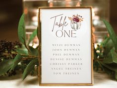 a table sign sitting on top of a table next to green leaves and flowers with candles in the background