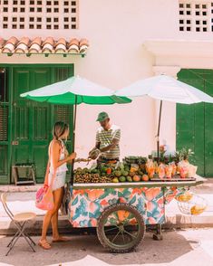 a man and woman standing next to a table with fruit on it under umbrellas