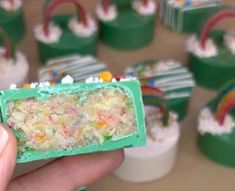 a hand holding a piece of cake in front of cupcakes with rainbow decorations on them