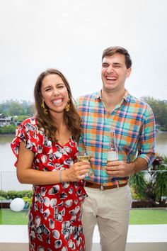 a man and woman standing next to each other holding wine glasses in front of a body of water