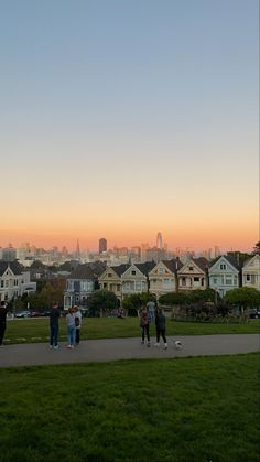 some people are standing in the grass and one is flying a kite with buildings in the background