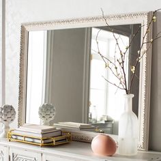 a white dresser topped with a mirror next to a vase filled with flowers and books