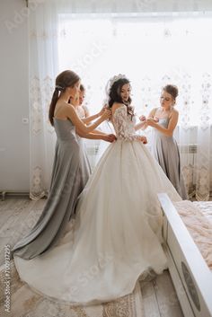 three bridesmaids helping the bride put on her wedding dress in front of a window