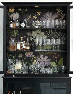 a black cabinet with floral wallpaper and wine glasses on the top shelf is filled with liquor