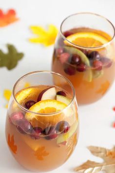 two glasses filled with fruit and ice on top of a white table next to leaves