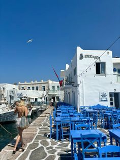 a woman is walking down the sidewalk near some blue tables and chairs in front of a white building