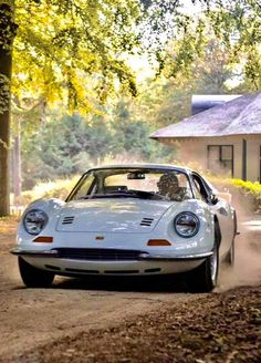 a white car driving down a dirt road next to a tree filled forest and a house