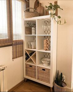 a white bookcase with wicker baskets and plants in it next to a window