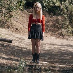 a woman with long blonde hair wearing a red shirt and black skirt standing in the woods