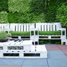 a wooden bench sitting next to a white table on top of a stone floor in front of a lush green wall