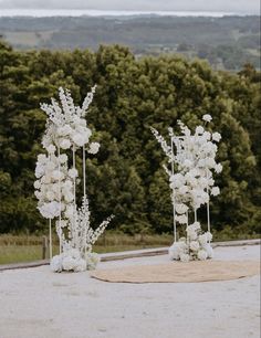 two tall white flower arrangements sitting on top of a stone slab in front of trees