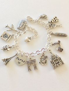 "Adorable Paris charm bracelet featuring 13 detailed silver charms of travel to Paris, classic landmarks, artist quarter, food and drink. Hand stamped 1/2\" 18 aluminum disc says \"Paris ❤️\". Memories of a visit abroad living \"la bonne vie\" in the magical city of Paris! Charms include: suitcase, plane, passport, fleur de lis, Eiffel Tower, Arc de Triumph, artist palette, Sacre Coeur church, Vespa scooter, baguette, cheese, wine, and croissant. Charms are antique silver, pewter and silver tone Silver Charms Jewelry Souvenir, Nickel-free Silver Charms Souvenir, Nickel-free Silver Charms For Souvenir, Silver Metal Jewelry For Travel, Silver Symbolic Jewelry Souvenir, Silver Engraved Jewelry For Travel, Silver Nickel-free Bracelet Souvenir, Nickel-free Silver Bracelets As Souvenir, Silver Charm Bracelet As Souvenir