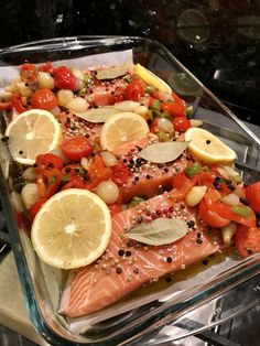 a glass dish filled with salmon, tomatoes, onions and lemons on top of a wooden table