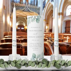 two white candles sitting on top of a table in front of a church pews