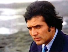 a man wearing a suit and tie looking off into the distance with water in the background