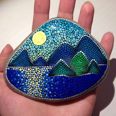 a hand holding a painted rock with mountains in the background and a moon on top