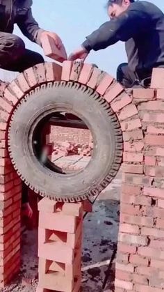 two men are building a brick wall with an old tire on the top and another man is holding something in his hand