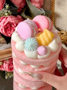 a hand holding a pink cake with white frosting and decorations on it, surrounded by flowers