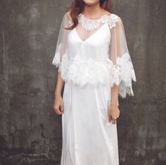 a woman standing in front of a gray wall wearing a white dress with sheer sleeves