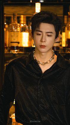 a young man wearing a black shirt and gold necklace standing in front of liquor bottles