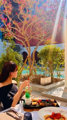 a woman sitting at a table with food in front of her and a tree behind her