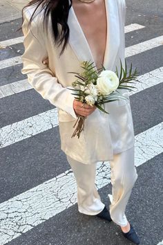 a woman in a white suit holding a bouquet of flowers while standing on the street