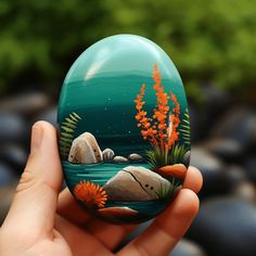 a hand holding up a painted rock with flowers and rocks in the water behind it