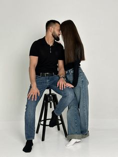 a man and woman sitting on top of a wooden stool next to each other in front of a white wall