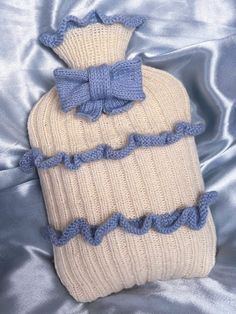 a white and blue knitted hot water bottle with a bow on the top sitting on a silver blanket